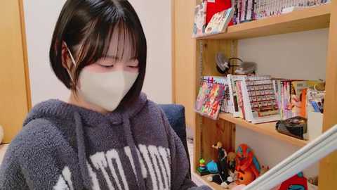 Video of an Asian woman with short black hair wearing a gray hoodie and mask, sitting at a desk cluttered with toys and books, surrounded by a cozy, cluttered room.