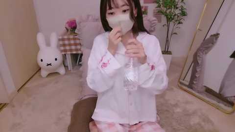 Media: A young Asian woman with shoulder-length black hair, wearing a white bunny-themed pajama set, kneels on a beige carpet, holding a mask in front of a full-length mirror, in a cozy, softly lit room with a white rabbit toy and potted plant in the background.