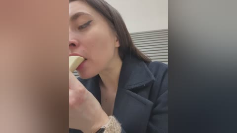 Video of a fair-skinned woman with straight brown hair, wearing a dark blazer, licking an apple while seated in a modern office setting with neutral-colored walls and horizontal blinds.