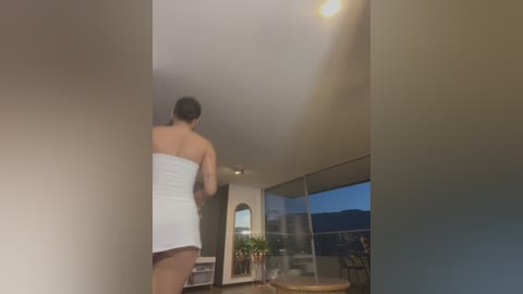 Media: Video of a woman with long dark hair, wearing a white towel, looking out a large window at a sunset over mountains from a modern, minimalist living room.