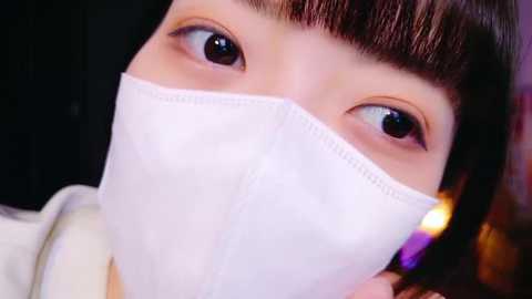 Media: A close-up video of an East Asian woman with light skin, wearing a white surgical mask covering her nose and mouth. She has dark brown hair with bangs and is gazing intently.