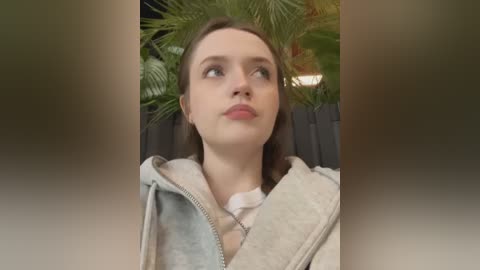 Media: Video of a young Caucasian woman with fair skin, brown hair, and blue eyes, wearing a grey hoodie, looking upward with a thoughtful expression, surrounded by lush green palm fronds.