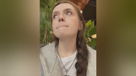 Media: Video of a young woman with light skin and long brown hair in braids, wearing a beige cardigan and a silver necklace, gazing upward with a serene expression. Lush green foliage and a brown hat are in the background.