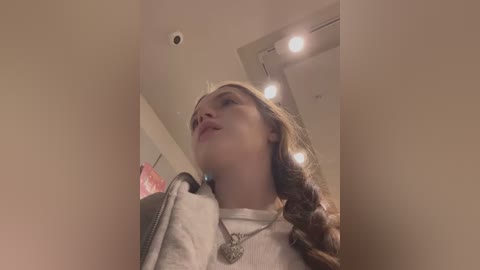 Media: Video of a young woman with long brown hair in a white shirt, looking upwards, taken from a low angle. She wears a silver necklace with a heart pendant.