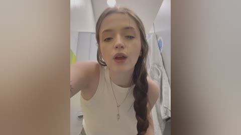 Media: Video of a young woman with fair skin, long brown hair in a braid, wearing a white tank top, leaning forward in a modern, brightly lit bathroom with white walls.