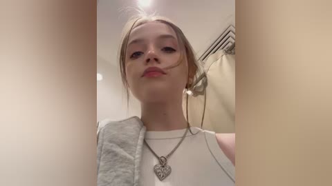 Media: A video of a young woman with fair skin, light brown hair, and light makeup, wearing a white sleeveless top and a silver heart necklace, in a bathroom with beige walls and a towel rack.