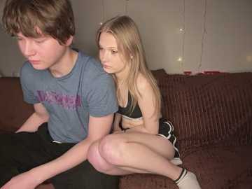 Video of a young couple, a boy with short hair in a blue shirt, and a girl with long blonde hair in a black sports bra and shorts, sitting on a brown couch.