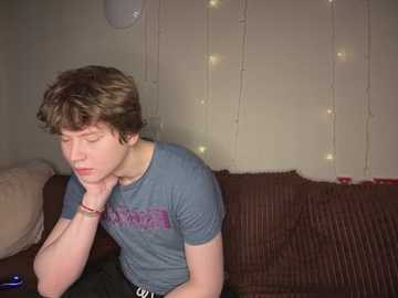 Video of a young man with short brown hair, wearing a blue T-shirt, resting his head on his hand, sitting on a brown couch with beige cushions.