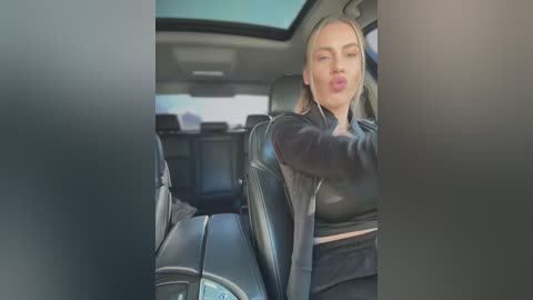 Media: Video of a blonde woman with light skin, wearing a black leather jacket and black pants, leaning back in a car seat with headphones on, making a pouty face.