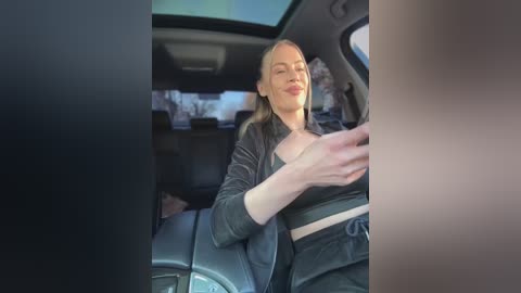 Media: Video of a blonde woman with fair skin, wearing a black jacket and bralette, seated in a car, smiling, with a blurred background.