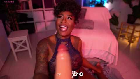 Video of a joyful Black woman with short curly hair and tattoos, wearing a blue lace halter top, sitting on a white chair in a dimly lit room with pink and purple lighting.