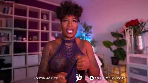 Media: A video of a confident Black woman with an afro, wearing a sheer, dark purple halter top, standing in a vibrant, modern room with bookshelves and flowers.