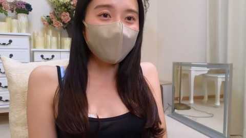 Media: Video of a young Asian woman with long black hair, wearing a beige face mask and black tank top, in a cozy, floral-decorated room with white furniture and a glass table.