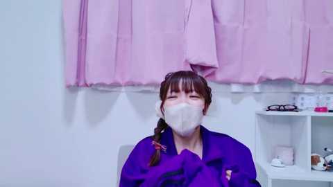 Media: Video of a woman in a purple coat, wearing a white surgical mask, sitting in a white room with pink curtains and a white shelf holding medical supplies.