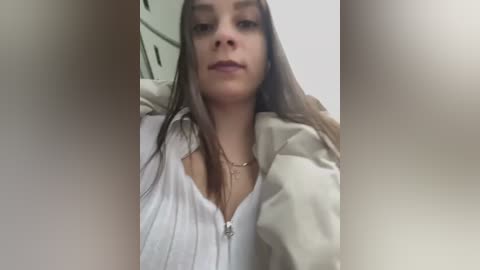 Media: Video of a young Caucasian woman with long, straight brown hair, fair skin, and a slender build, wearing a white blouse and beige jacket, seated indoors with blurred background.