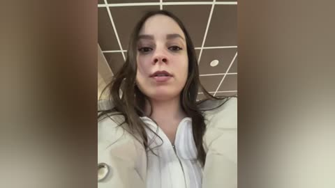 Media: Video of a young woman with long brown hair, wearing a white shirt, looking straight into the camera, in an office setting with a grid ceiling.