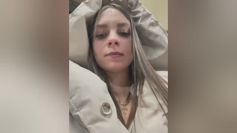 Media: Video of a young woman with light skin and long, straight, gray hair, wearing a beige, button-up jacket, lying on a bed with white pillows. The background is blurred, focusing on her relaxed, neutral expression.