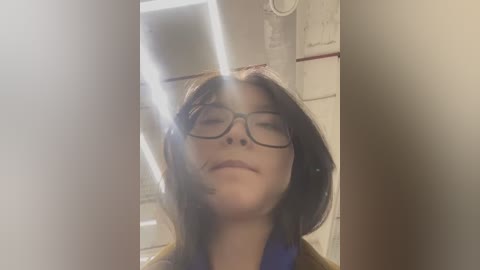 Media: Video of a woman with long brown hair, wearing black-rimmed glasses, smiling. She stands in a dimly lit bathroom with white tiles and a shower curtain.