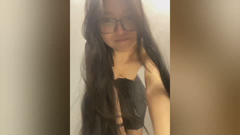 Media: Video of a young woman with long, dark hair, wearing glasses, black top, and plaid scarf, standing against a beige wall.