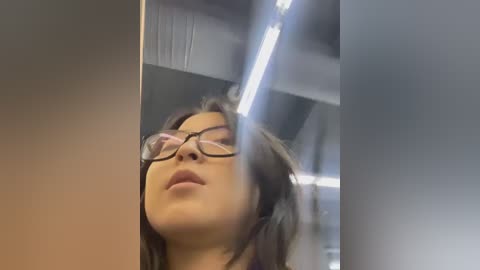 Media: Video of an Asian woman with long, dark hair and glasses, looking upwards with a neutral expression, taken in a modern indoor setting with white ceiling panels and fluorescent lighting.