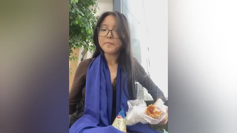 Media: A video of an Asian woman with glasses, long black hair, wearing a blue robe, eating takeout food in a modern indoor setting.