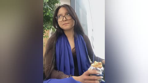 Media: A video shows a young woman with long brown hair, glasses, and a blue scarf, eating a sandwich, standing indoors beside a potted plant.