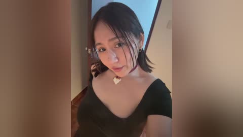 Media: A video of an Asian woman with short dark hair, wearing a black top, standing in a hallway with beige walls. She has a small heart necklace.