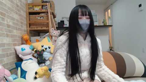 Media: Video of a woman with long black hair, wearing a white mask and a beige sweater, seated on a couch with stuffed animals and a wooden shelf in the background.