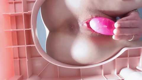 Video of a woman's exposed genitalia with a pink vibrator inserted, positioned in a pink plastic laundry basket. The background is a grid of pink and white squares.