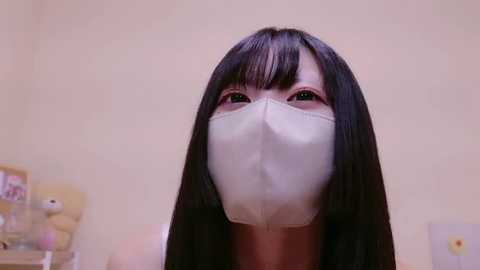 Media: Video of a woman with long black hair wearing a white face mask, standing indoors against a beige wall. The background features a framed picture and a shelf with various items.