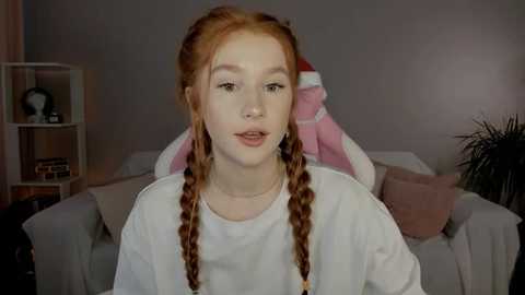 Media: Video of a young woman with pale skin, red braided hair, and a white sweater. She sits on a gray couch in a cozy living room with pink and brown cushions.