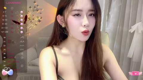 Media: Video of an East Asian woman with long, straight brown hair, fair skin, wearing a black spaghetti strap top, sitting in a well-lit room with sheer white curtains.