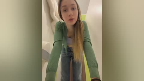 Media: Video of a young Caucasian girl with straight, light brown hair, wearing a green cardigan over a white top and blue jeans. She leans forward, looking directly at the camera. The background features a beige wall and a white towel hanging.