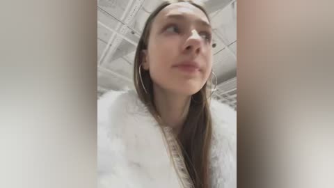 Media: A video of a young Caucasian woman with straight, brown hair, wearing a white fur-trimmed jacket, standing in a tiled room with white walls and a ceiling. She has a slight smile and is gazing upward.