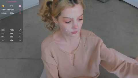 Media: A video of a young woman with light skin, blonde hair, and freckles, wearing a beige shirt, sitting on a gray floor, looking down, in a modern indoor setting.