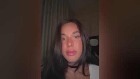 A video of a young woman with light brown skin and straight, shoulder-length dark brown hair. She has a neutral expression, wearing a black choker necklace and a white top. The background shows a dimly lit room with dark curtains.