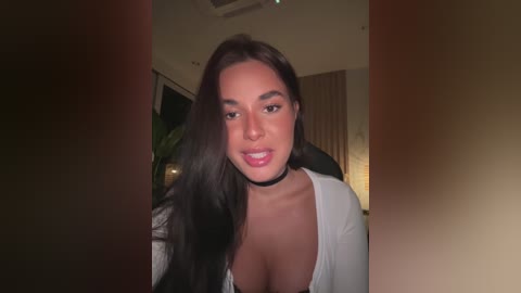 Media: Video of a young woman with long, dark hair and fair skin, wearing a white top and black choker, smiling warmly in a dimly lit room.