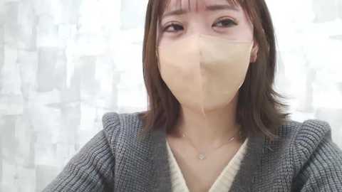 Media: A video of a young woman with short, dark brown hair and light skin, wearing a gray checkered blazer over a white top, and a clear plastic face mask. She has a calm expression. The background is a white curtain with abstract patterns.