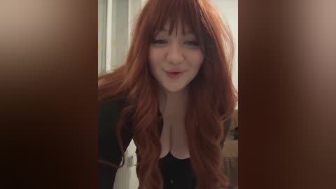 Media: A video of a young woman with long, wavy red hair, fair skin, and a black top, smiling in a bathroom with brown-tiled walls and a towel rack in the background.