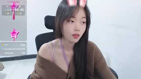 Media: Video of an East Asian woman with long black hair, wearing a brown jacket and a pink lace bra, seated on a black chair in a minimalist room.