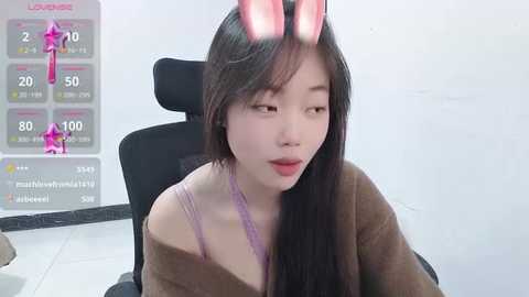 Media: Video of a young East Asian woman with long black hair, wearing a pink bunny ears headband, a purple lace bra, and a brown cardigan, seated in a black office chair.