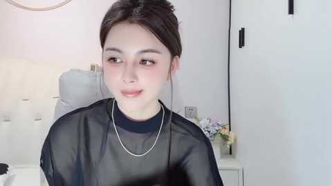 Media: Video of a young East Asian woman with fair skin, brown hair, and rosy cheeks, wearing a black sheer top and silver necklace. She sits in a modern, minimalist room with white walls and a bed in the background.