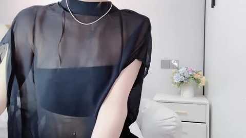 A video of a fair-skinned woman wearing a sheer black blouse with a high neckline, a silver necklace, and a white bed in the background with a vase of pastel flowers.