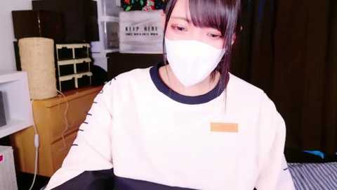 Media: Video of an Asian woman with straight black hair, wearing a white face mask and a white long-sleeve shirt with a yellow patch, sitting in a cluttered room with a TV, lamp, and cardboard boxes.