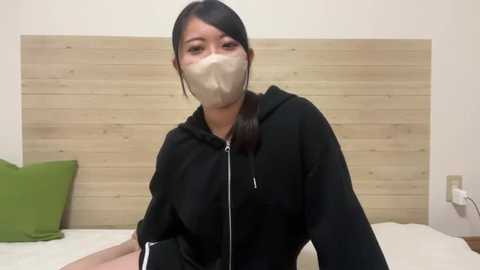 Video of an Asian woman with long black hair and a beige surgical mask, wearing a black hoodie, sitting on a bed with a light wooden headboard and green pillow.