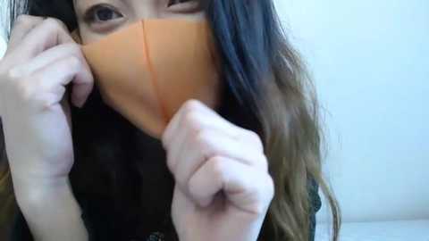 Media: Video of a young woman with long, dark hair and light skin, wearing an orange face mask, smiling with raised fists, against a plain white background.