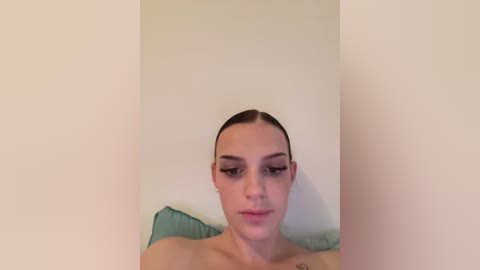 Media: A video of a fair-skinned woman with slicked-back hair, wearing minimal makeup and a green pillow, set against a plain white wall.