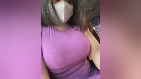 Media: A video of a woman with a light brown complexion wearing a white face mask, a purple ribbed top, and a black shoulder bag. She has straight, shoulder-length dark hair. The background is blurred in soft pink hues.