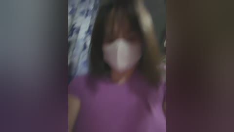 Media: A blurred video of a person with long brown hair, wearing a purple shirt and a white face mask, standing in a dimly lit room with a blue floral patterned wall.