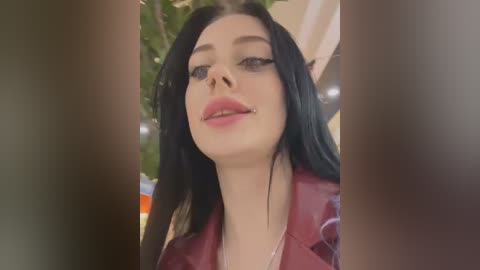 Media: Video of a young woman with pale skin, long black hair, and red lipstick, wearing a maroon leather jacket, indoors with blurred background.